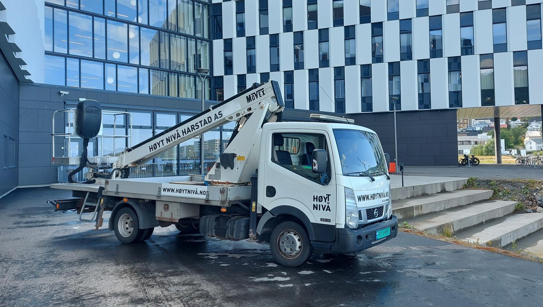 Liftbil fra Høyt Nivå Harstad parkert utenfor moderne kontorbygg med store glassfasader, klar for arbeid på vinduer og fasade.